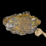 A rock embedded with intricate circuit board patterns, held delicately by pale hands drawn in a ghostly style. The contrast between the rough, metallic mineral and the sleek, artificial circuit board illustrates the relationship between raw natural resources and modern technological development. The hands evoke human involvement in the extraction and manufacturing processes.